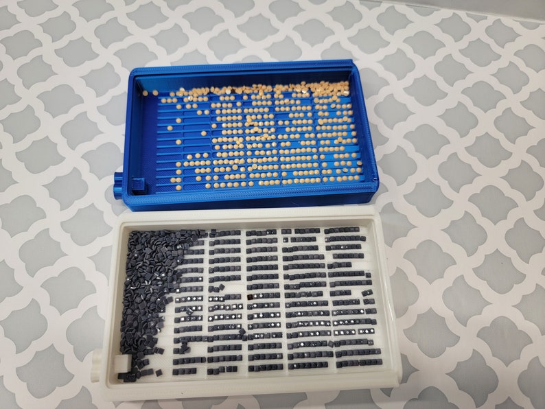 Puede incluir: Dos bandejas de pl&aacute;stico para organizar los taladros de pintura de diamantes. La bandeja superior es azul y tiene peque&ntilde;os taladros redondos de color beige. La bandeja inferior es blanca y tiene filas de peque&ntilde;os taladros cuadrados negros.