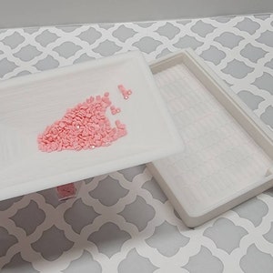May include: A white plastic tray with a small pile of pink diamond-shaped rhinestones in the center. The tray is part of a two-piece set, and the other tray is visible in the background.
