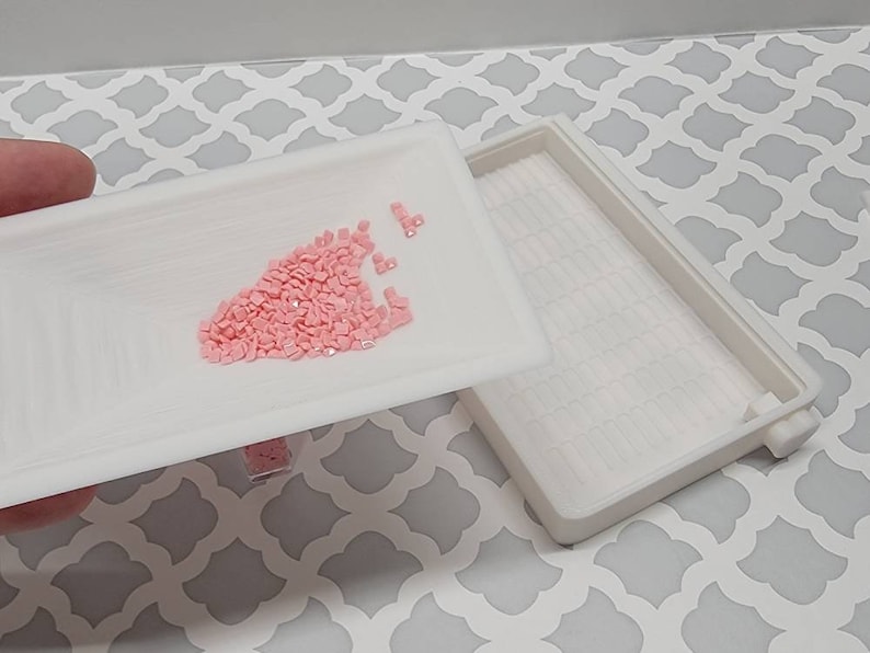 May include: A white plastic tray with a small pile of pink diamond-shaped rhinestones in the center. The tray is part of a two-piece set, and the other tray is visible in the background.
