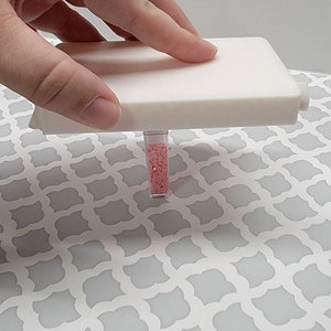 May include: A white plastic container with a clear tube filled with pink diamond-shaped beads. The container is open and the tube is visible.