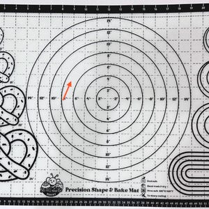 May include: A white baking mat with a grid pattern and a circle with concentric circles in the center. The mat has a variety of shapes printed on it, including pretzels and spirals. The text "Precision Shape & Bake Mat" is printed on the mat.