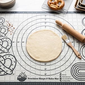 May include: A rectangular silicone baking mat with black printed guides for pretzels, circles, and spirals. The mat features a ruler border in inches and centimeters, a wooden rolling pin, a pastry brush, and a round piece of raw dough.