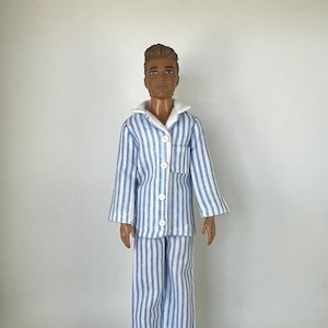 May include: A doll wearing blue and white striped pajamas. The pajamas include a button-down shirt with a chest pocket and matching pants. The doll has brown hair and is standing on a clear plastic stand.