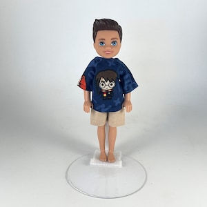 May include: A doll wearing a navy blue t-shirt with a Harry Potter graphic and khaki shorts. The t-shirt has a pattern of blue shapes. The doll is standing on a clear plastic stand.