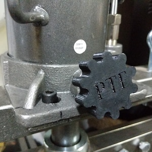 May include: Close-up of a black plastic gear knob with the letters "PTF" embossed on it. The knob is attached to a gray metal machine.