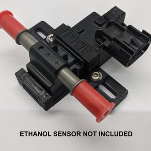 Puede incluir: Componente automotriz de plástico negro con extremos rojos y tubos plateados. El componente tiene una base rectangular y un conector negro. El texto "ETHANOL SENSOR NOT INCLUDED" está en la parte inferior.