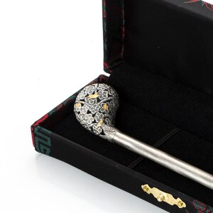 May include: A silver and gold decorative stick with a detailed floral design on the top, resting in a black box with a red and green floral pattern. The box has the text "NASCENKA" on it.