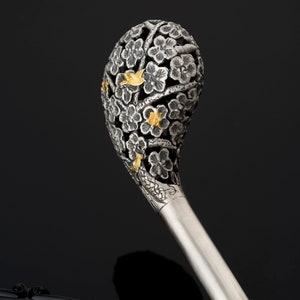 May include: A silver hair stick with a decorative top featuring a floral design with gold accents. The top is shaped like a sphere and is intricately carved with flowers and leaves.