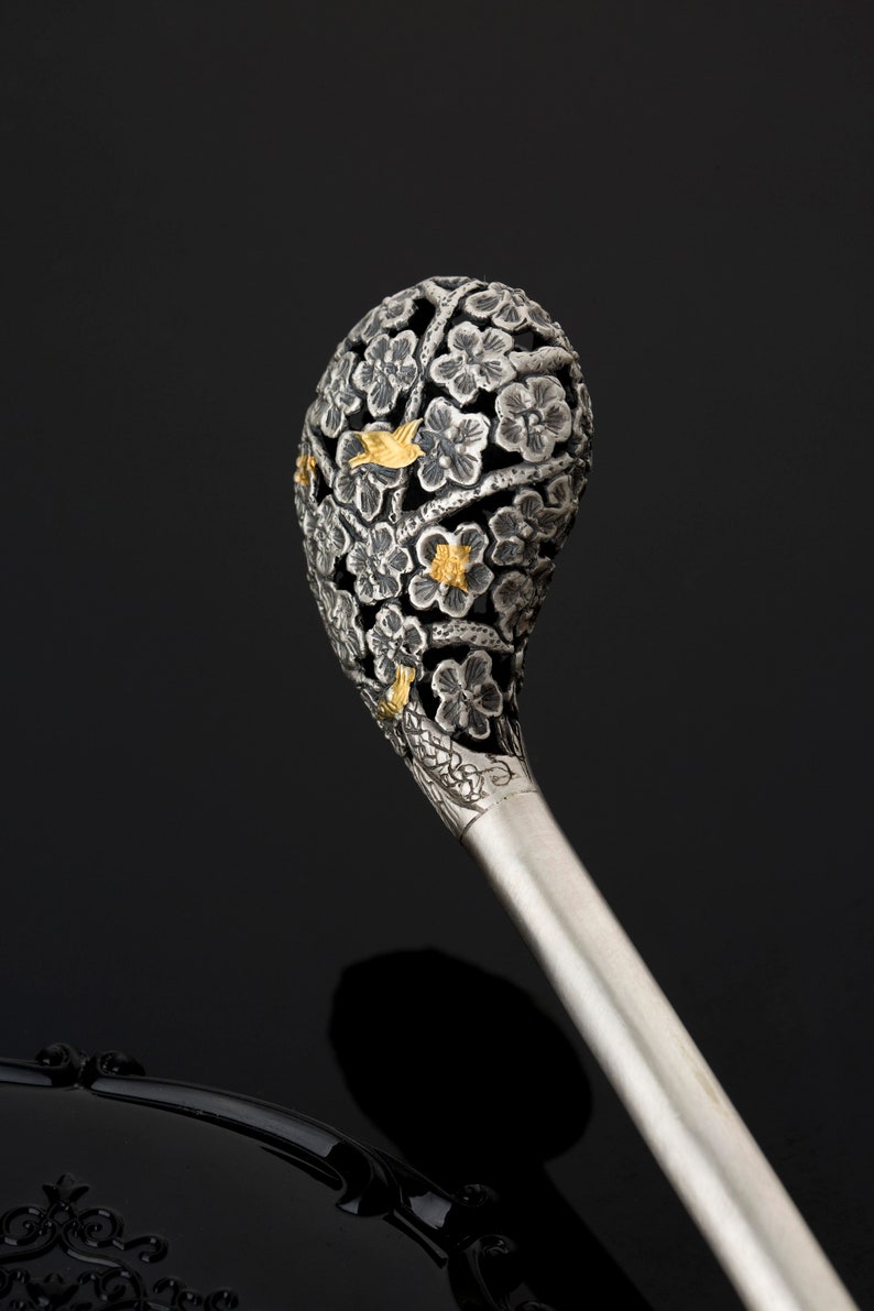 May include: A silver hair stick with a decorative top featuring a floral design with gold accents. The top is shaped like a sphere and is intricately carved with flowers and leaves.