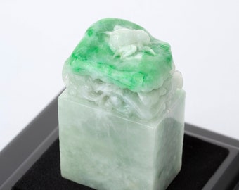 Puede incluir: Un sello de jade verde con tallas intrincadas, que presenta un diseño de dragón y una pieza de jade más pequeña en la parte superior. El sello se muestra sobre una base negra.