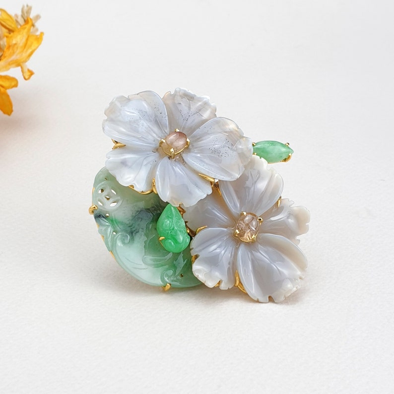以下が含まれることがあります： 緑色の翡翠の葉と金のアクセントが付いた白い翡翠の花のブローチ。ブローチはピンク色のセンターを持つ白い花が2つあり、緑色の翡翠のベースにセットされています。