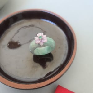 May include: A jade ring with a light green hue, adorned with a delicate, light pink flower and a small green gem at its center. The ring is displayed on a dark brown, glossy surface.