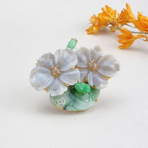 以下が含まれることがあります： 2つの白い花と緑の葉を持つ、翡翠と貝殻の花のブローチ。花は金色のアクセントと中央にピンクの宝石が付いています。