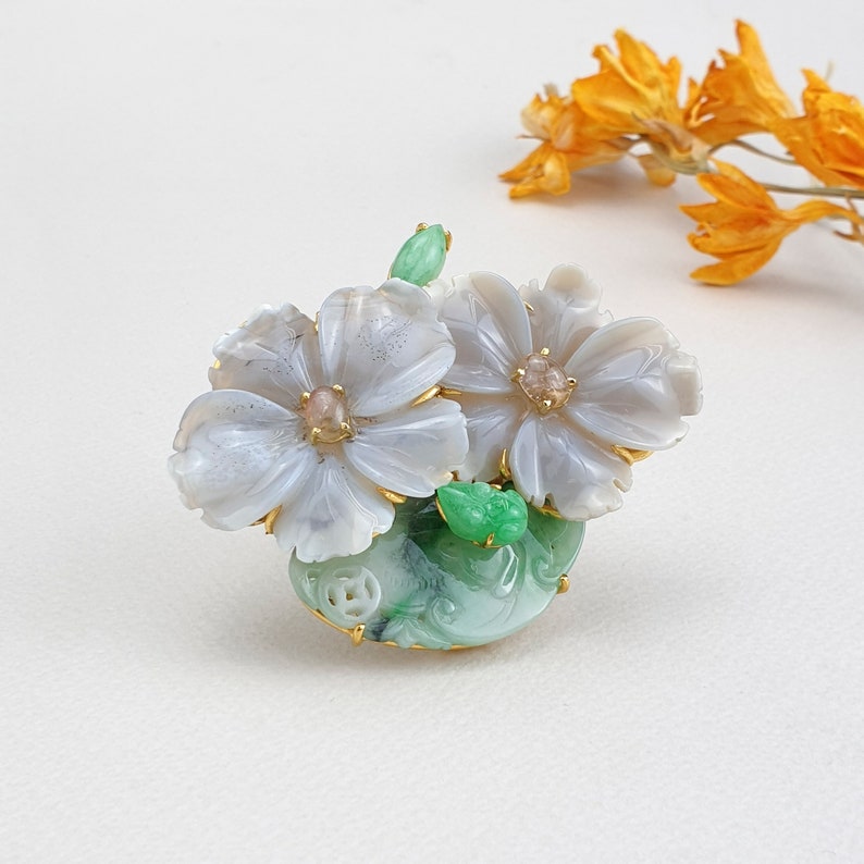 以下が含まれることがあります： 2つの白い花と緑の葉を持つ、翡翠と貝殻の花のブローチ。花は金色のアクセントと中央にピンクの宝石が付いています。