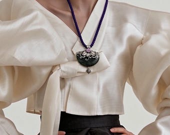 May include: A cream-colored wrap blouse with voluminous sleeves and a tie-front closure, paired with a black skirt. A dark purple cord necklace features a silver pendant with a dark green stone, a light pink accent, and a small silver drop charm.