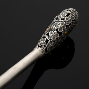 May include: A silver and gold decorative stick with a floral design. The stick is made of metal and has a long, thin handle. The top of the stick is decorated with a cluster of small flowers.
