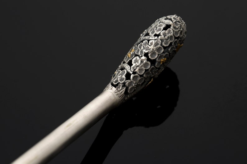 May include: A silver and gold decorative stick with a floral design. The stick is made of metal and has a long, thin handle. The top of the stick is decorated with a cluster of small flowers.