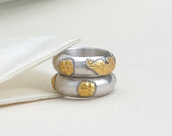 Puede incluir: Dos anillos de plata con detalles dorados. Cada anillo presenta un diseño dorado martillado y un patrón dorado decorativo. Los anillos están apilados, mostrando los intrincados detalles de la joyería.