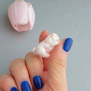 Peut inclure: Une main avec un vernis &agrave; ongles bleu paillet&eacute; tient une petite figurine de cochon en bois blanc. Le cochon est couch&eacute; sur le c&ocirc;t&eacute;, la t&ecirc;te reposant sur ses pattes avant.