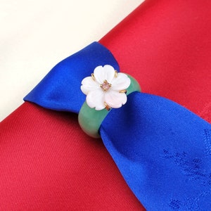 Può includere: Un anello porta tovagliolo in giada verde con un designo a forma di fiore bianco e una pietra preziosa rosa al centro. L'anello è su uno sfondo di tessuto blu e rosso.