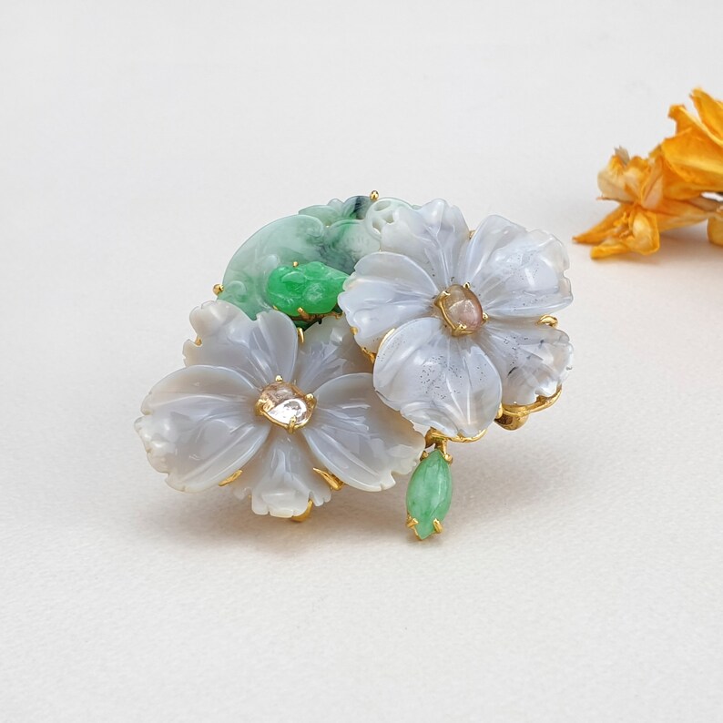 以下が含まれることがあります： 白とグレーの瑪瑙で彫られた3つの花が特徴の繊細なブローチ。緑色の翡翠の葉とクリアなラインストーンでアクセントが付けられています。ブローチはゴールドトーンの金属でセットされています。
