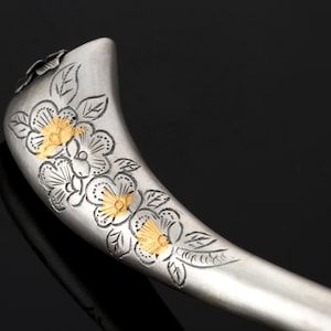 May include: Silver hair stick with a floral design. The flowers are engraved with gold accents.
