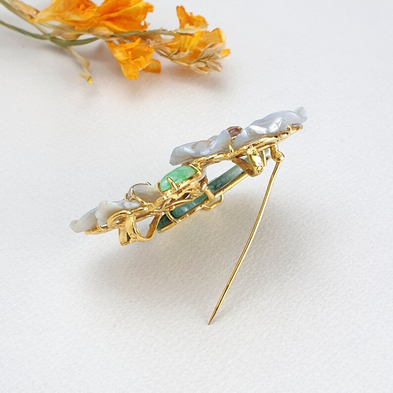 以下が含まれることがあります： 真珠と緑色の石のデザインが施された金のブローチ。ブローチの裏側にピンが付いています。
