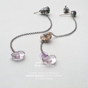 May include: Elegant silver earrings with a dangling chain design. Each earring features a small, ornate stud, a delicate flower, and a teardrop-shaped, light purple gemstone. The earrings are set against a white background.