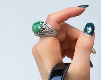 Könnte beinhalten: Ein silberner Ring mit einem grünen Edelstein und floralem Design. Der Ring befindet sich an einem Finger, und die Nägel der Person sind grün lackiert.