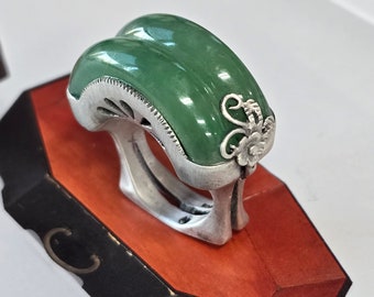 May include: A silver ring with a green jade stone and intricate silver detailing. The ring is set on a red and black display stand.