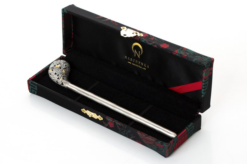 May include: A silver and gold decorative stick with a detailed floral design on the top, resting in a black box with a red and green floral pattern. The box has the text "NASCENKA" on it.