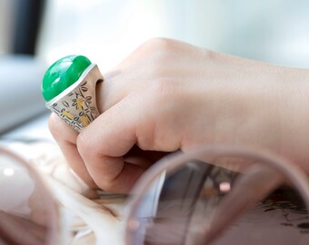 Könnte beinhalten: Ein silberner Ring mit einem großen, grünen Edelstein und filigranen Blumenmustern. Der Ring liegt auf einem weißen Hintergrund.