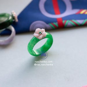 May include: A vibrant green jade ring with a delicate pink flower accent and a small green gemstone center. The ring is set against a light background with a patterned fabric. The website naschenka.com and Instagram handle @nas.naschenka are visible.