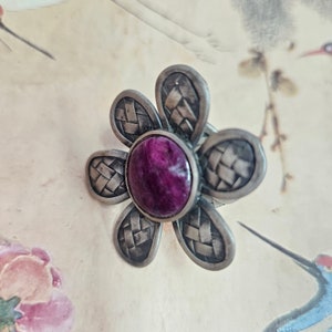 May include: A silver flower-shaped ring with a purple gemstone in the center. The ring has a woven design on the petals.