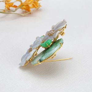 以下が含まれることがあります： 白と緑の翡翠の石を使った花柄の金色のブローチ。ブローチは衣服に付けるためのピンが後ろについています。