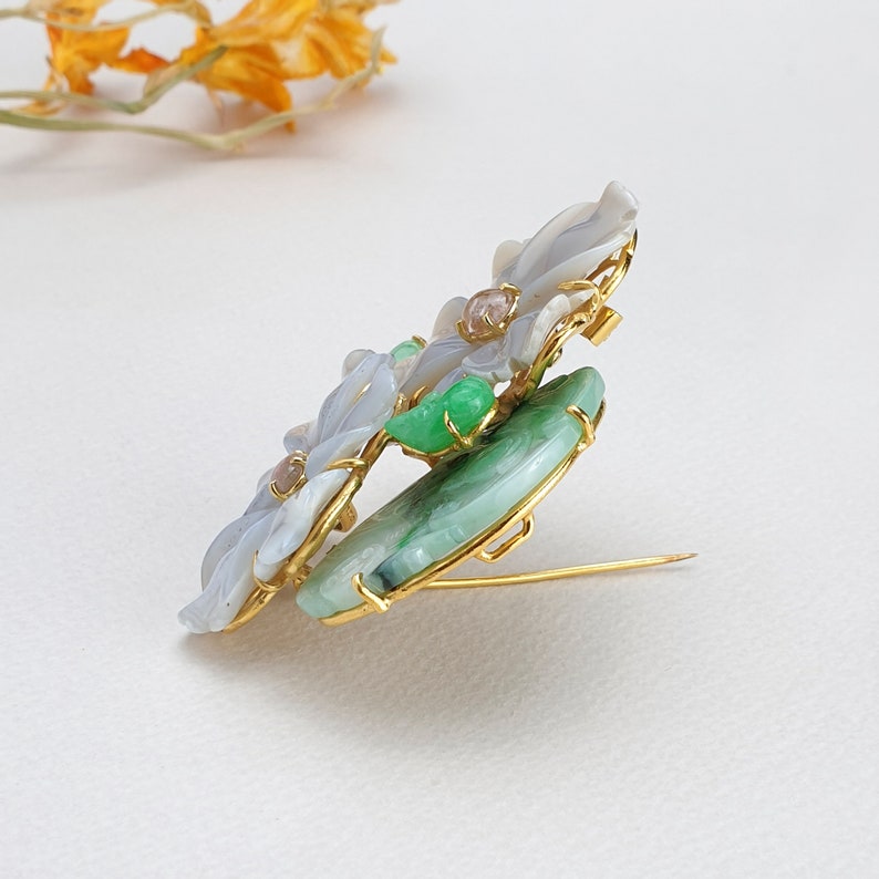 以下が含まれることがあります： 白と緑の翡翠の石を使った花柄の金色のブローチ。ブローチは衣服に付けるためのピンが後ろについています。