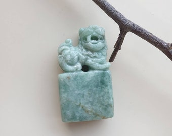 Puede incluir: Una talla de jade verde de un león mítico, también conocido como foo dog, sentado sobre una base cuadrada. La talla es detallada e intrincada, con un diseño chino tradicional.