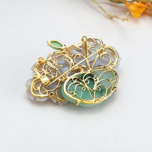 以下が含まれることがあります： 中央に緑色の翡翠の石がついた金のフィリグリーペンダント。ペンダントは、複雑なディテールが施された花の形をしています。翡翠の石は金の枠で囲まれています。