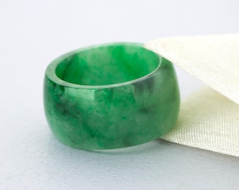 May include: A vibrant green jade ring with a polished finish. The ring has a thick band and a smooth, rounded interior. The jade displays natural variations in color, with darker green and black inclusions. The ring rests on a white surface.