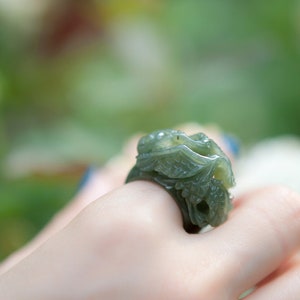 May include: A green jade ring carved with intricate floral designs. The ring is worn on a person's finger.