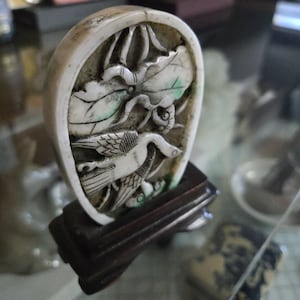 May include: An oval-shaped, off-white carving of a bird and foliage, mounted on a dark wooden stand. The carving features a detailed relief of a bird with outstretched wings and leaves with green accents. The stand is rectangular.