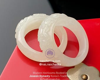 May include: Two white jade bangles with floral carvings, displayed on a red surface. The image includes text: naschenka.com, @nas.naschenka, and "Modern Heirlooms Rooted in Joseon Dynasty Korea's Tradition & Craftsmanship."
