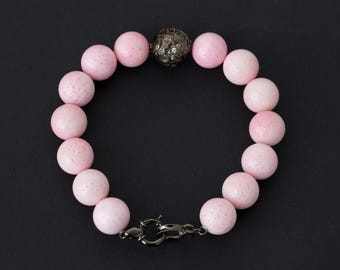 Puede incluir: Una pulsera con grandes cuentas redondas de color rosa claro. Las cuentas están conectadas por un cierre de metal oscuro y una cuenta decorativa. La pulsera se muestra sobre una superficie negra.