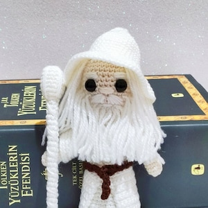 May include: White crocheted amigurumi figure of Gandalf the Grey from The Lord of the Rings. The figure is holding a white staff and wearing a brown belt. The figure is sitting on a book with the title "Yüzüklerin Efendisi" in Turkish.