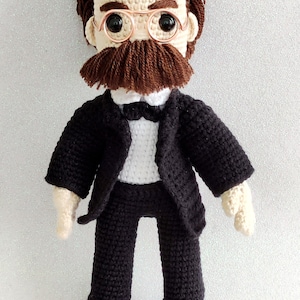 May include: A crocheted doll wearing a black suit, white shirt, and bow tie. The doll has brown hair, a brown moustache, and is wearing round glasses.