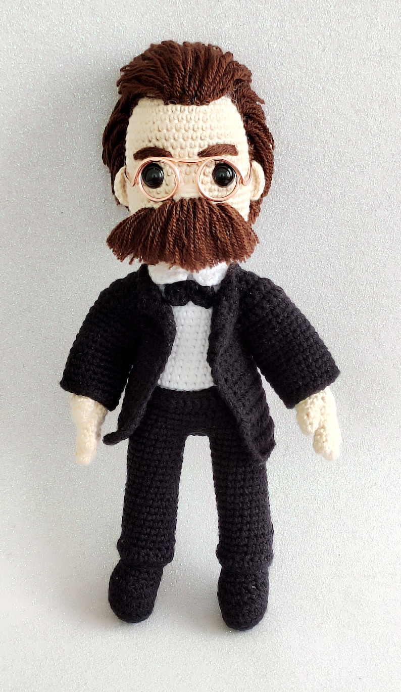 May include: A crocheted doll wearing a black suit, white shirt, and bow tie. The doll has brown hair, a brown moustache, and is wearing round glasses.