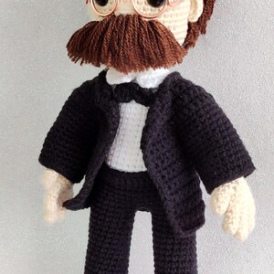 May include: Crocheted doll wearing a black suit with a white shirt and bow tie. The doll has brown hair, a brown moustache, and is wearing round, gold-rimmed glasses.