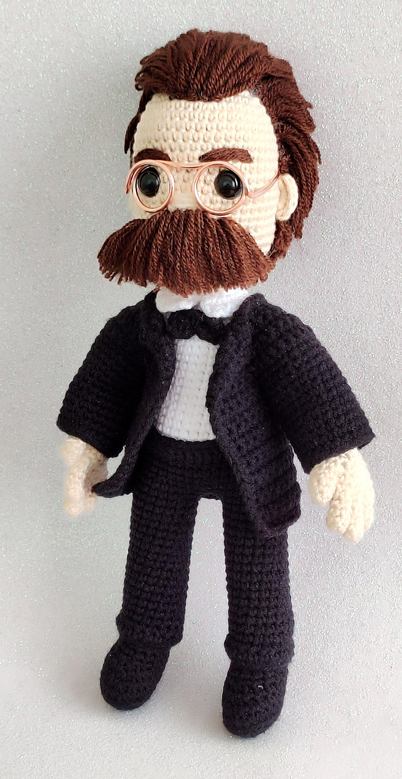 May include: Crocheted doll wearing a black suit with a white shirt and bow tie. The doll has brown hair, a brown moustache, and is wearing round, gold-rimmed glasses.