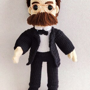 May include: Crocheted doll wearing a black suit, white shirt, and bow tie. The doll has brown hair, a moustache, and round glasses.