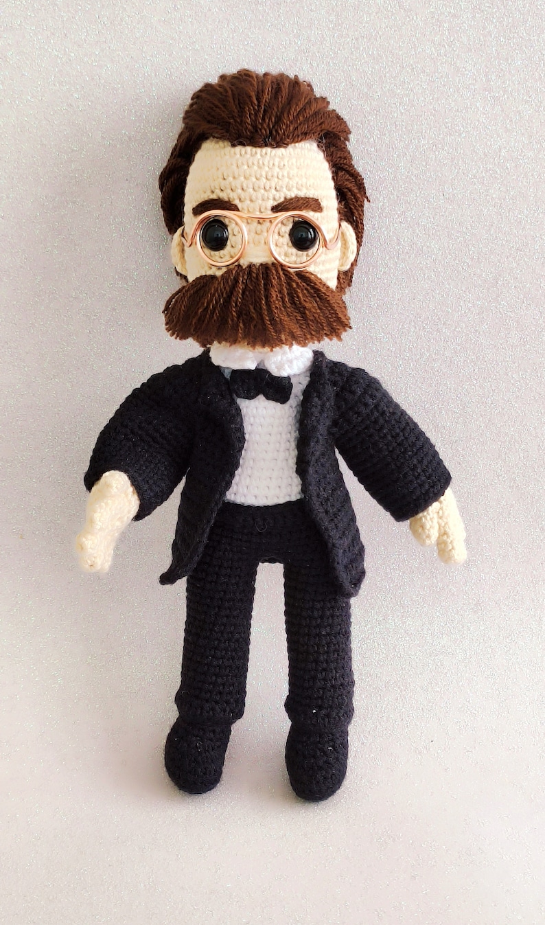 May include: Crocheted doll wearing a black suit, white shirt, and bow tie. The doll has brown hair, a moustache, and round glasses.